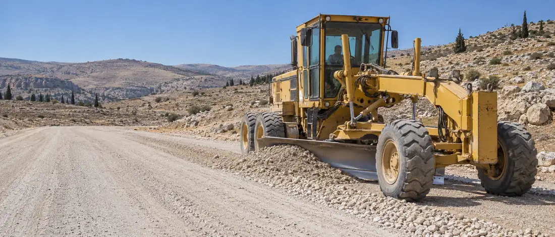 A motor grader spreading and levelling a gravel layer between rocky hills