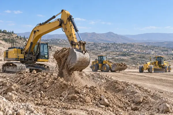 An excavator dumping a bucket of soil, with a loader and a grader behind it on an earthworks site