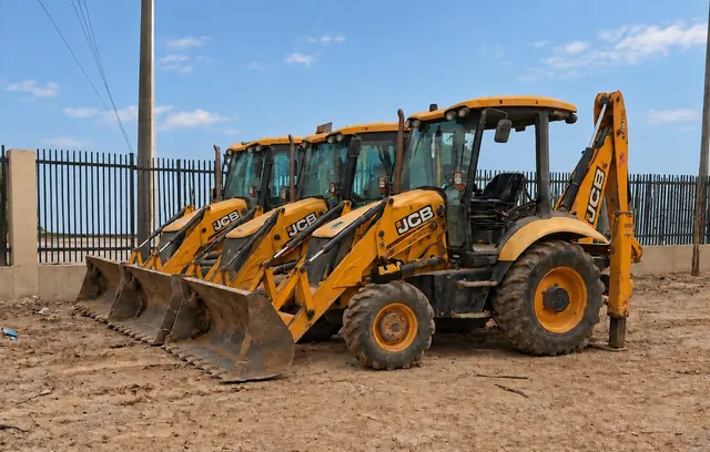 Three backhoe loaders lined up in a fenced dirt yard, their front buckets resting on the ground