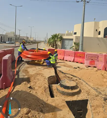 Workers laying orange cable ducts in a trench next to a manhole, on a site fenced off with barriers