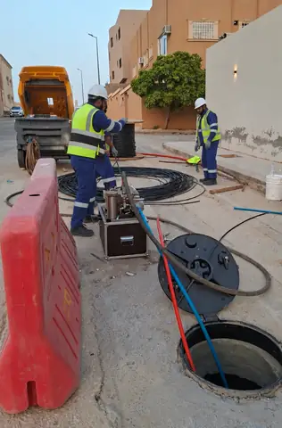 Technicians beside an open manhole blowing fiber optic cable into its ducts with a blowing machine, with the cable drum behind them