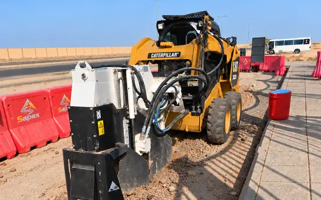 A narrow-trench cutter opening a cable route along the edge of a street next to the curb, with Sapex-branded safety barriers enclosing the work site